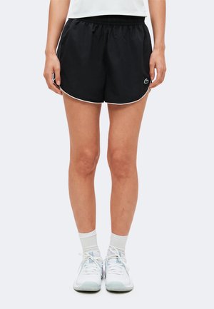 Personne portant un short de sport noir avec un liseré blanc, des chaussettes blanches et des baskets blanches, debout devant un fond uni.