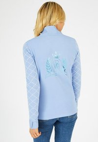 Haut bleu clair à manches longues avec un col haut, arborant un emblème décoratif et un motif en diamants sur les manches.