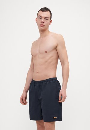 Navy swim shorts featuring an elastic waistband, side pockets, and a small orange logo detail on the left leg. Smooth fabric texture.