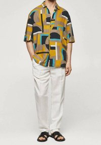 Camisa de manga corta con un patrón geométrico en colores mostaza, negro, marfil y azul verdoso, combinada con pantalones blancos y sandalias negras.