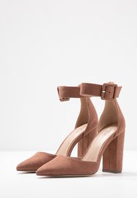 Brown suede high-heeled pumps with a pointed toe, ankle strap, and a chunky heel. Smooth texture with a minimalistic design.