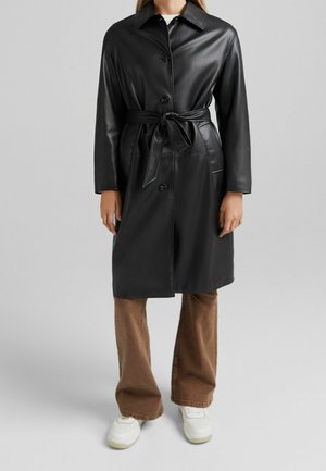 Femme portant un manteau en simili cuir noir ceinturé, boutonné devant, un pantalon évasé marron et des baskets blanches, debout sur un fond gris.