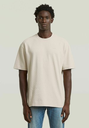 Jonge man met kort krullend haar die een loszittend beige textuur T-shirt en een blauwe spijkerbroek draagt, staand tegen een lichte achtergrond.