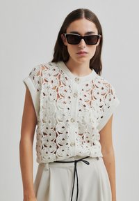 Top croșetat alb cu un model floral, mâneci scurte și deschidere cu butoane. Asortat cu o fustă plisată de culoare deschisă și ochelari de soare negri.
