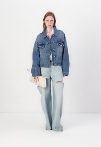 Veste en denim avec deux poches avant, finie dans un délavage bleu moyen, assortie à un jean à jambes larges bleu clair avec des déchirures subtiles. Tenant une pochette beige.