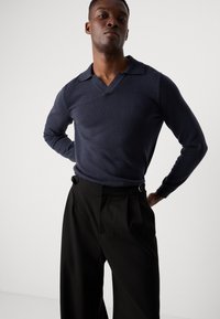 Jersey de punto texturizado en azul marino con escote en V y cuello amplio, combinado con pantalones negros con plisados que cuentan con herrajes laterales ajustables.