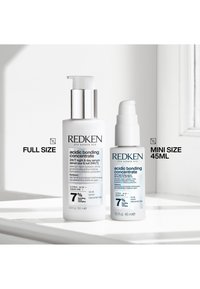 Twee Redken zure bonding concentrate serumflessen met etiketten vol formaat 100 ml en mini formaat 45 ml op een wit oppervlak bij een raam.