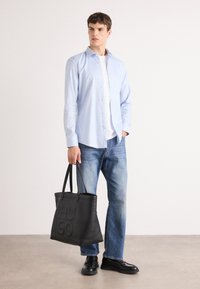 Bolso tote negro con logotipo "HUGO" en relieve, textura suave, sostenido por un modelo, combinado con una camisa de rayas azul claro y jeans de mezclilla.