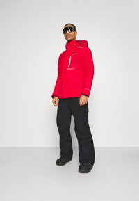 Röd vattentålig jacka med justerbar huva och framficka, i kombination med svarta cargobyxor och svarta vandringsskor. Modellen står mot en grå bakgrund.