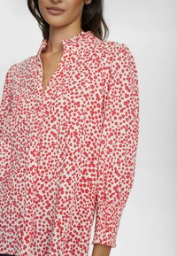 Blusa bianca con stampa floreale rossa, caratterizzata da scollo a V, patta con bottoni e polsini elasticizzati. Il materiale sembra leggero con una finitura testurizzata.