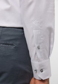 Weißes, strukturiertes langärmliges Hemd mit einem knöpfbaren Ärmel, ausgestattet mit zwei Metallknöpfen und einem klaren, strukturierten Kragen, kombiniert mit grauen Hosen.