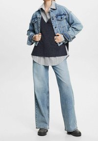 Jeansjacke, dunkler Strick-Weste und gestreiftes Hemd darunter. Hellblaues, weites Jeansmodell, schwarze Schuhe, lässige Passform, minimale Hardware.