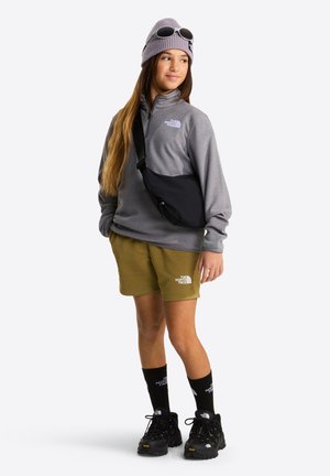 Pulover gri din fleece, pantaloni scurți verde măsliniu, geantă de umăr neagră, pantofi de drumeție negri și o căciuliță tricotată mov cu ochelari de protecție, stând în fața unui fundal alb.