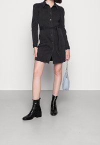 Robe en denim noir avec manches longues, boutonnière à l'avant et ceinture. Associée à des bottines noires et un petit sac bandoulière bleu clair.