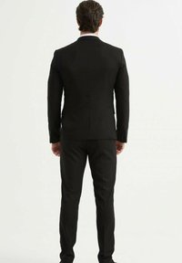 Veste de costume noire avec revers en pointe, assortie à un pantalon ajusté. Le tissu semble lisse avec une silhouette cintrée et un éclat subtil.