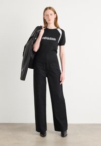 Juoda trumpa rankovė marškinė su balta užrašu "calvin klein", derinama su plačiais juodais kelnais, kurie turi subtilų blizgesį. Modelis laiko juodą odinę striukę.