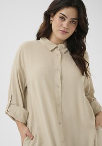 Beige knoopshirt van zachte stof, met een kraag, lange mouwen met opgerolde tabs en zijzakken. Eenvoudig ontwerp met een relaxte pasvorm.