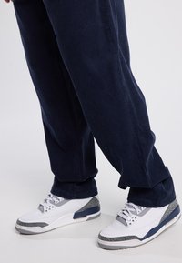 Meripihkaiset samettihousut, joissa on pystyribbaus, yhdistettyinä valkoisiin lenkkitossuihin, joissa on harmaat yksityiskohdat ja karkeat kuvioidut pohjat.