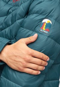 Niebieska kurtka puchowa o pikowanej fakturze. Posiada haftowaną naszywkę z logo w żywych kolorach przedstawiającą drzewa i góry na rękawie.