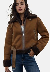 Veste en shearling marron avec un col en fourrure, fermeture éclair sur le devant, poches latérales et accents en cuir. Comprend une taille ceinturée et des poignets texturés.