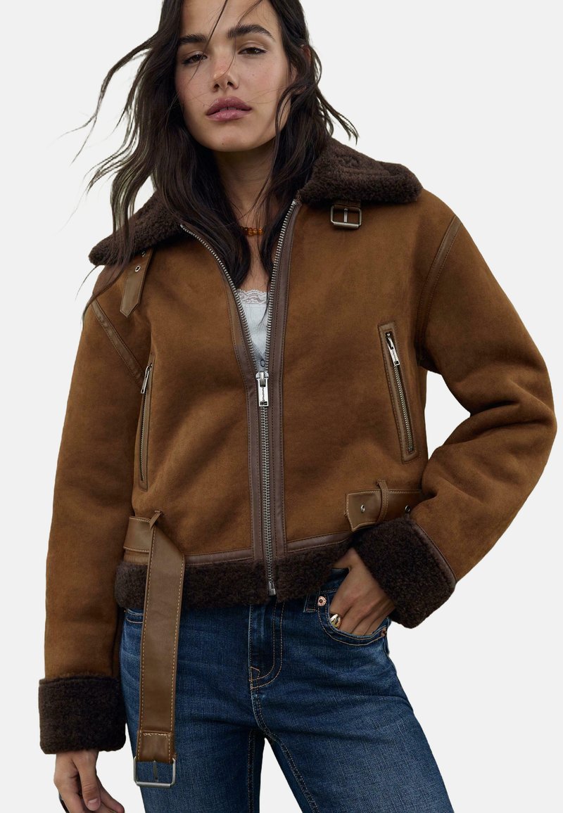 Veste en shearling marron avec un col en fourrure, fermeture éclair sur le devant, poches latérales et accents en cuir. Comprend une taille ceinturée et des poignets texturés.
