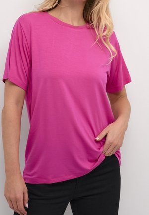 Kobieta ubrana w jasnoróżowy krótki rękaw T-shirt w połączeniu z czarnymi spodniami, trzymająca dół koszulki na jednolitym jasnym tle.