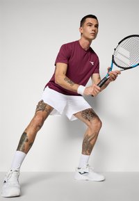 Atleta con tatuaggi che indossa una maglietta borgogna, pantaloni corti bianchi e sneaker, tiene una racchetta da tennis in una postura attiva e pronta.