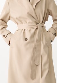 Beżowy trencz z paskiem, podwójnym zapięciem z przodu i dwoma kieszeniami. Posiada klasyczny kołnierz i akcenty w postaci guzików. Gładka tekstura tkaniny.