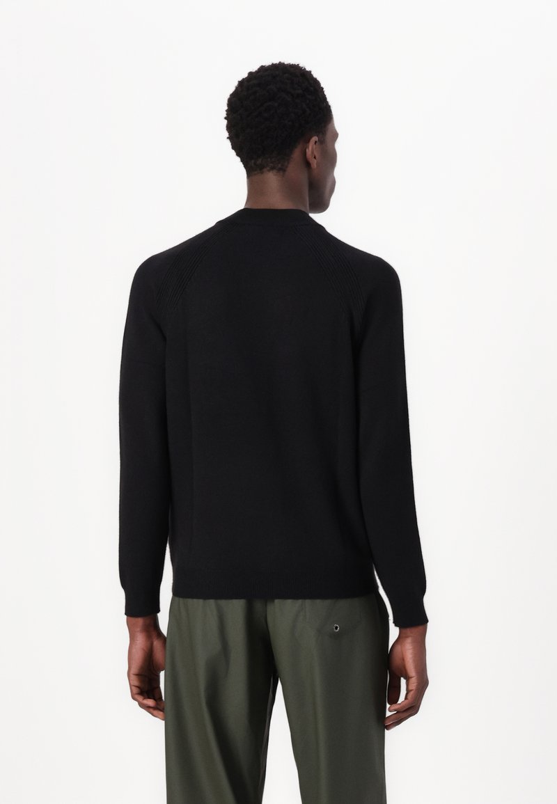 Schwarzer Pullover mit Rundhalsausschnitt, gerippten Akzenten an den Schultern und langen Ärmeln, kombiniert mit grünen Hosen. Glatte Textur.