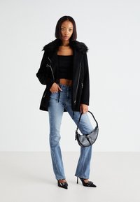 Čierny kožúšok s umelou kožušinou na čiernom crop topu, svetlo modré džínsy s postrannými ozdobami, čierne lodičky a textúrovaná kabelka.