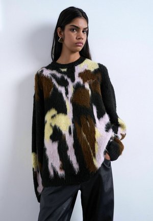 Giovane donna che indossa un maglione peloso con motivo astratto nero, marrone, giallo e rosa e pantaloni neri, su uno sfondo bianco uniforme.