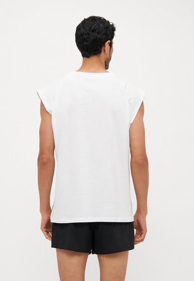 White sleeveless t-shirt made of cotton, featuring a round neck, loose fit, and visible stitching details. Worn with black shorts.