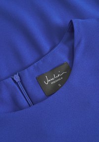 Modrá blúzka vyrobená z hladkej látky s okrúhlym výstrihom. Má stredný zips a čierny štítok s nápisom "Vashti STOCKHOLM S."