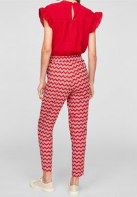 Blusa rossa con volant e schiena a chiave, abbinata a pantaloni con motivo zigzag in rosso e bianco, e sneakers di colore chiaro.