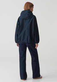 Marineblauwe waterdichte jas met capuchon, elastische manchetten en verstelbare zoom; gecombineerd met donkere jeans en casual schoeisel. Minimalistisch ontwerp.