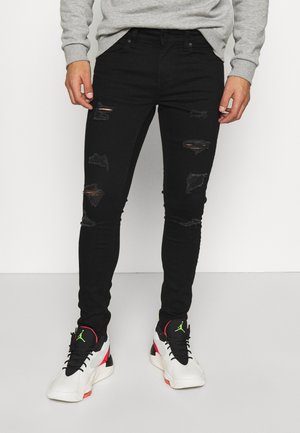 Mężczyzna ubrany w czarne postrzępione obcisłe dżinsy, białe i czarne sneakersy z zielonym logo oraz jasnoszarą koszulę z długim rękawem, stojący na białym tle.