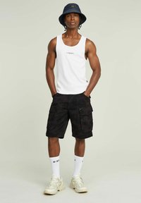 Biała bezrękawnik, czarne spodenki cargo w kamuflażu z zamkami błyskawicznymi, białe skarpetki, beżowe trampki i granatowy kapelusz typu bucket.