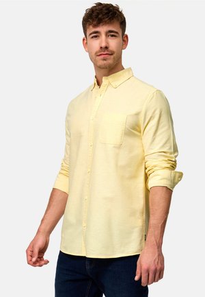 Ung mand iført en bleg gul skjorte med knapper og opkrogede ærmer samt mørke jeans, stående mod en ensartet lys baggrund.