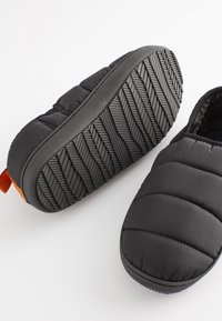 Chaussons matelassés noirs avec semelle en caoutchouc texturée et languette de tirage orange. Tige en tissu doux et rembourrage pour le confort.