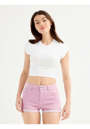 Joven mujer de pie mirando hacia adelante, lleva un top corto blanco y unos pantalones cortos de talle alto en rosa claro con bajo doblado.