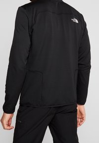Czarna polarowa kurtka z fakturowanym wykończeniem, długimi rękawami i bocznymi kieszeniami. Zawiera logo The North Face na górnej części pleców.