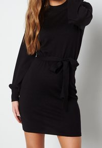 Robe en maille noire à manches longues avec une taille cintrée. Comprend une ceinture assortie à nouer et des poignets froncés sur les manches.