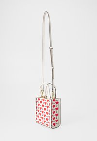 Štvorcová crossbody taška s bielym exteriérom so červenými pixelovanými srdci, zlatým hardvérom a krátkymi držadlami s dlhým nastaviteľným popruhom.