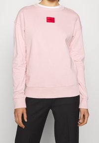 Sudadera rosa hecha de una tela suave, con cuello redondo, puños acanalados y un parche de logo bordado en rojo en el pecho.