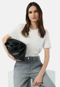 Schwarze Lederhandtasche mit einer weichen Textur, größerer runder Form, gehalten gegen ein schlichtes weißes Kurzarmhemd und graue hochgeschnittene Jeans.