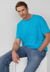 Türkises T-Shirt mit kurzen Ärmeln und Brusttasche, aus glattem Stoff gefertigt. Kombiniert mit hellblauen Jeans, sitzt das Model auf einem weißen Podest.