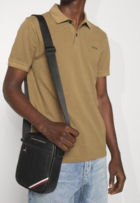 Polo marron foncé à deux boutons, associé à un jean bleu clair. Un sac bandoulière noir texturé avec un détail rayé rouge et blanc.