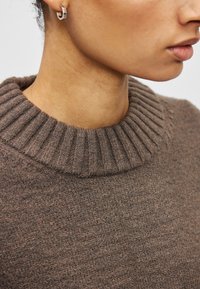 Καφέ πλεκτό πουλόβερ με ριμπ ύφασμα και ψηλό γιακά. Η κοντινή λήψη δείχνει το λείο ύφασμα με ένα κομψό, εφαρμοστό σχέδιο.