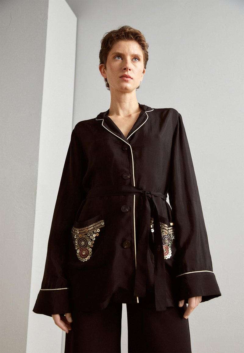Zwarte overhem shirt met lange mouwen, witte piping en geborduurde zakken met cirkelpatronen en pailletten. Strikdetail aan de taille.