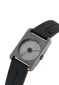 Montre carrée avec un boîtier en métal gris brossé, cadran gris, aiguilles noires et accent orange. Bracelet en caoutchouc noir au design épuré et avec un détail de logo.